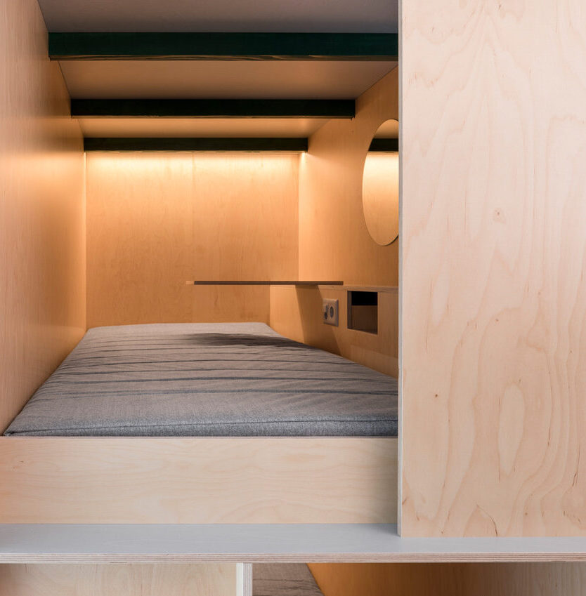 Blick in eine Schlafkapsel des Green Marmot Hotels mit grauer Matratze, indirekter Beleuchtung, aufgeklapptem Beistelltisch und rundem Spiegel an der Wand.