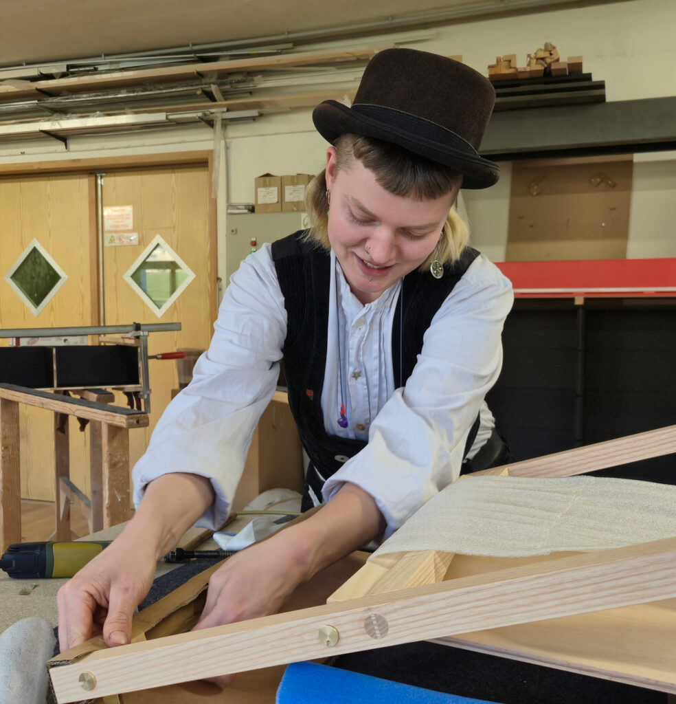 Wandergesellin mit schwarzem Hut und traditioneller schwarzer Weste über weißem Hemd arbeitet an einem Holzrahmen in einer Werkstatt