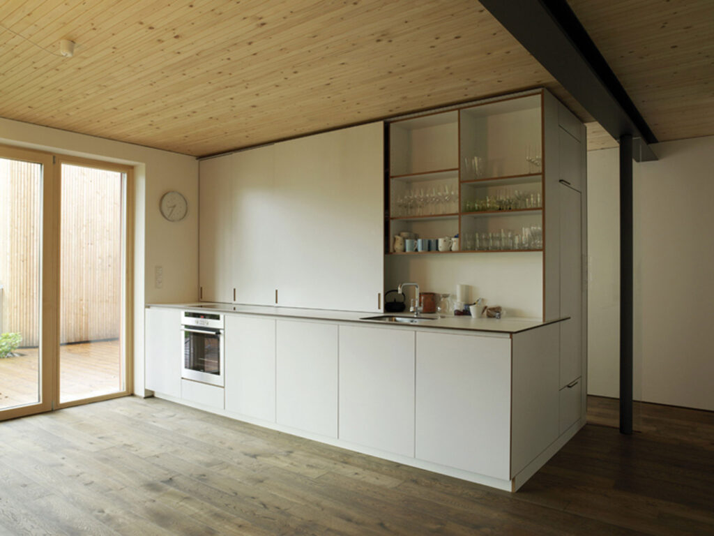 Moderne Einbauküche mit weißen Fronten, integriertem Backofen, offenem Regal mit Gläsern und Holzdecke in hellem Raum mit großem Fenster.