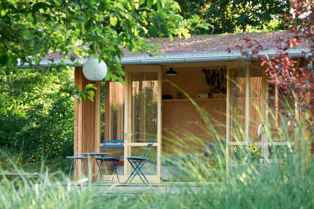 Holz-Gartenhaus mit großen Glasflächen, davor zwei Stühle und ein Tisch, umgeben von grüner Vegetation